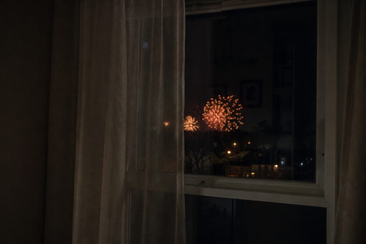 Distant fireworks seen through a closed window with the curtains partly drawn