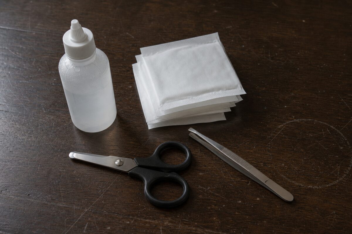 Sterile saline, antiseptic wipes, blunt scissors, and tweezers grouped on a scratched table